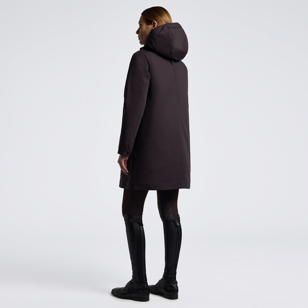 A woman shown from the back wearing a dark plum hooded coat, black leggings, and black boots against a clean light gray studio background.