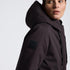 A close-up side profile of a model wearing a dark brown hooded jacket with a black rectangular logo patch on the upper sleeve, set against a plain white background.