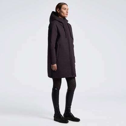 A model is shown in profile wearing a long, dark plum hooded parka jacket, styled with black patterned tights and black leather riding boots against a clean, light gray studio background.