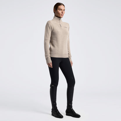 A woman modeling a beige ribbed knit mock-neck sweater with a small tonal logo on the left chest, paired with black fitted pants and black boots, against a light gray studio background.