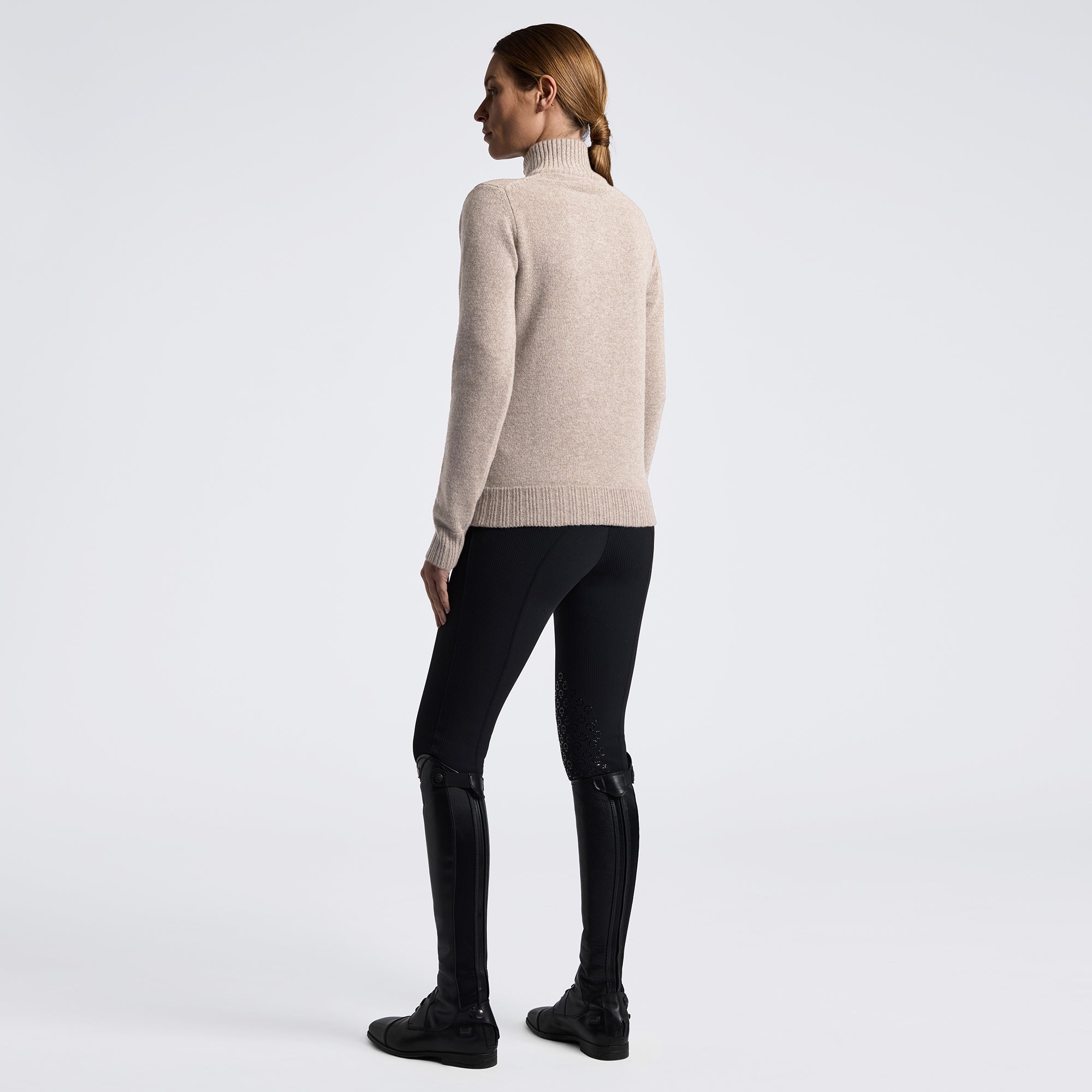 A woman shown from the back wearing a light beige turtleneck sweater, black fitted pants, and black riding boots against a plain light gray studio background.