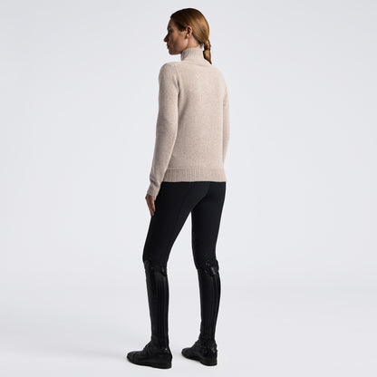 A woman shown from the back wearing a light beige turtleneck sweater, black fitted pants, and black riding boots against a plain light gray studio background.