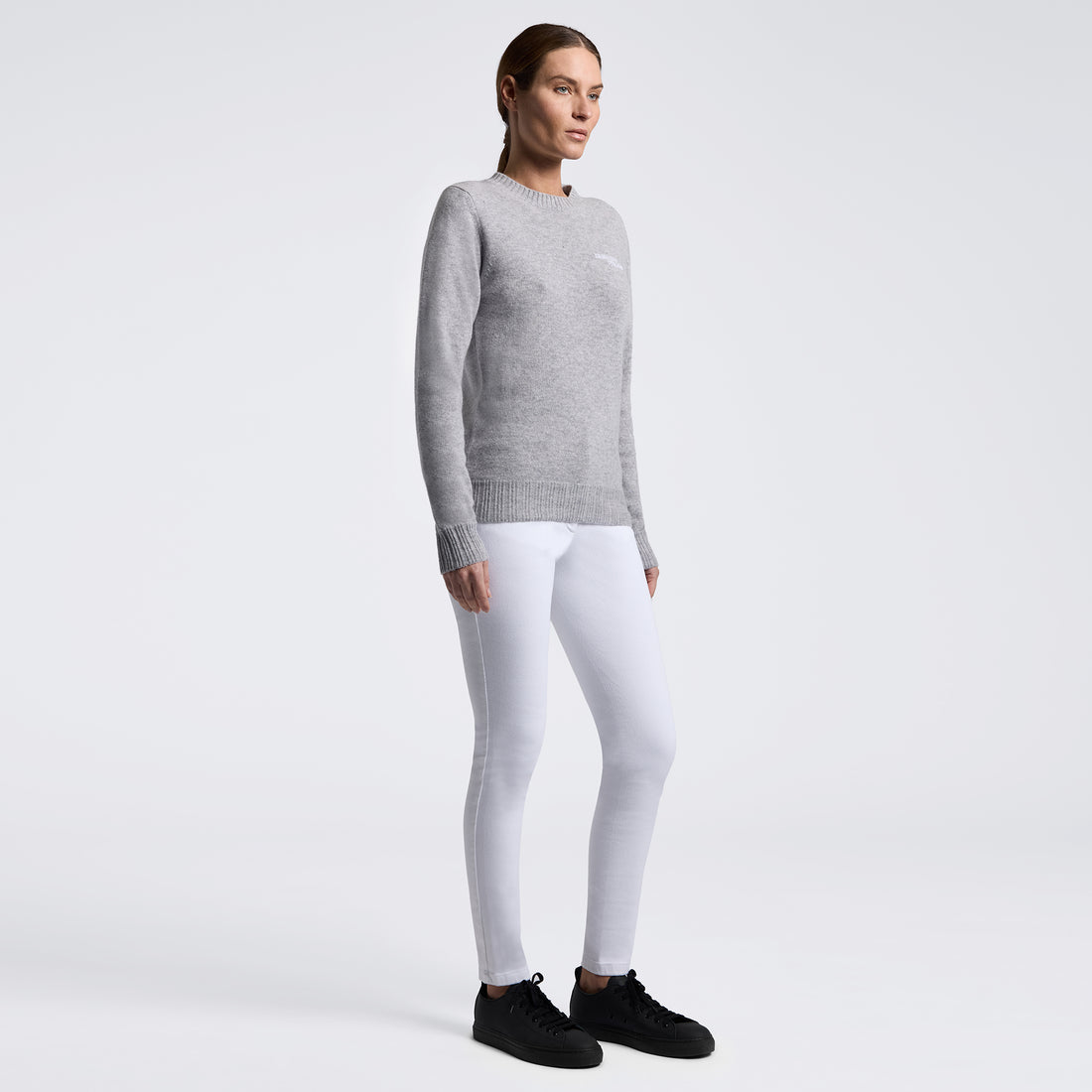 A woman modeling a light gray long-sleeve knit sweater and white leggings with black sneakers, standing against a plain light gray studio background.