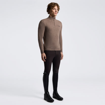 A male model wearing a brown long-sleeve quarter-zip knit top, black riding breeches with textured knee panels, and black riding boots, standing against a light gray studio background.
