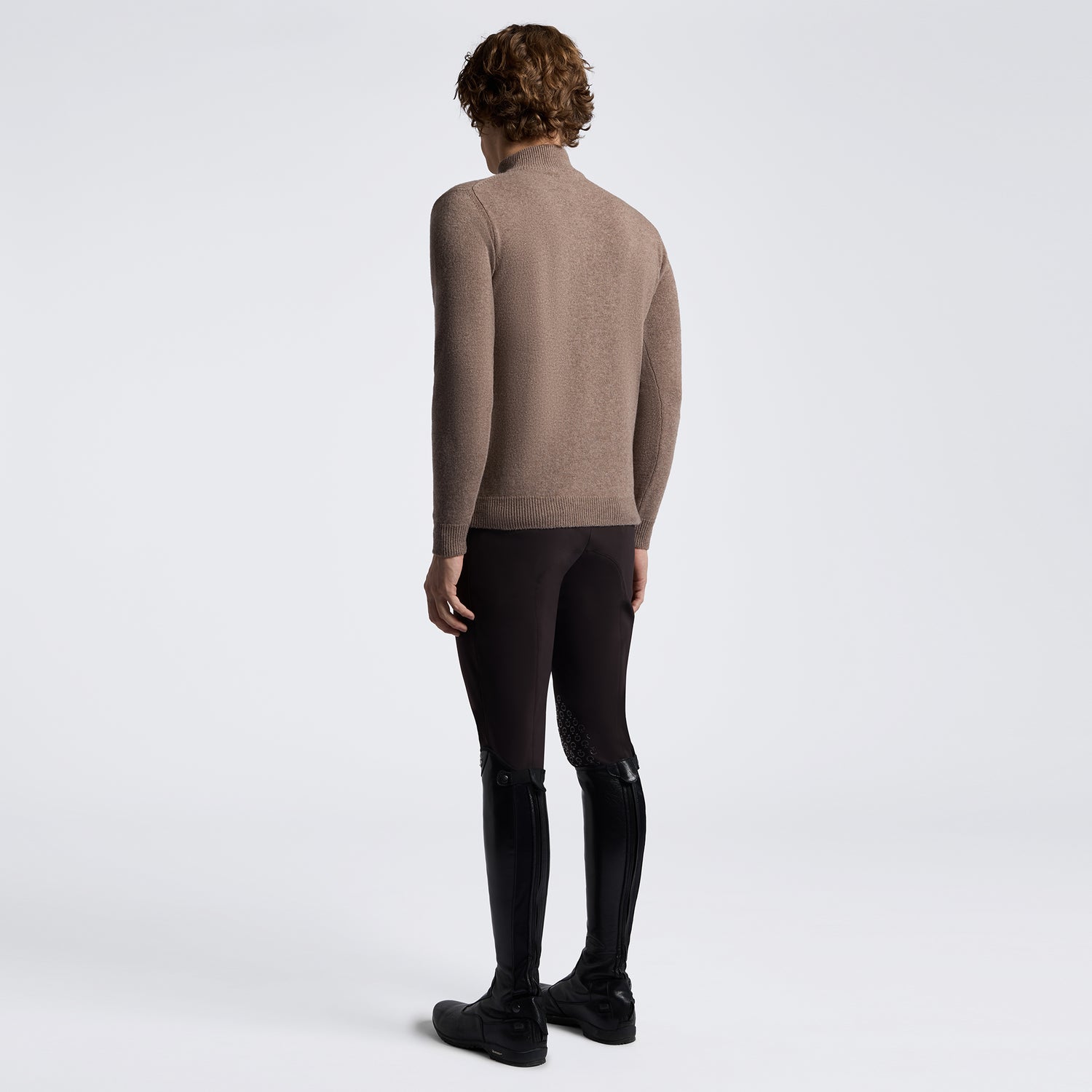 A model shown from the back wearing a taupe-brown long-sleeve mock neck sweater, black riding breeches, and black riding boots against a light gray studio background.