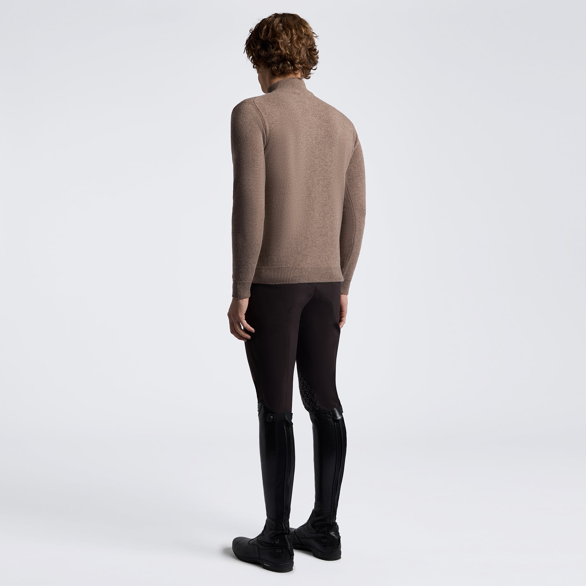 A model shown from the back wearing a taupe-brown long-sleeve mock neck sweater, black riding breeches, and black riding boots against a light gray studio background.