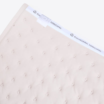 A close-up of a light beige baby blanket featuring a repeating anchor pattern and a white elastic band with the brand name &